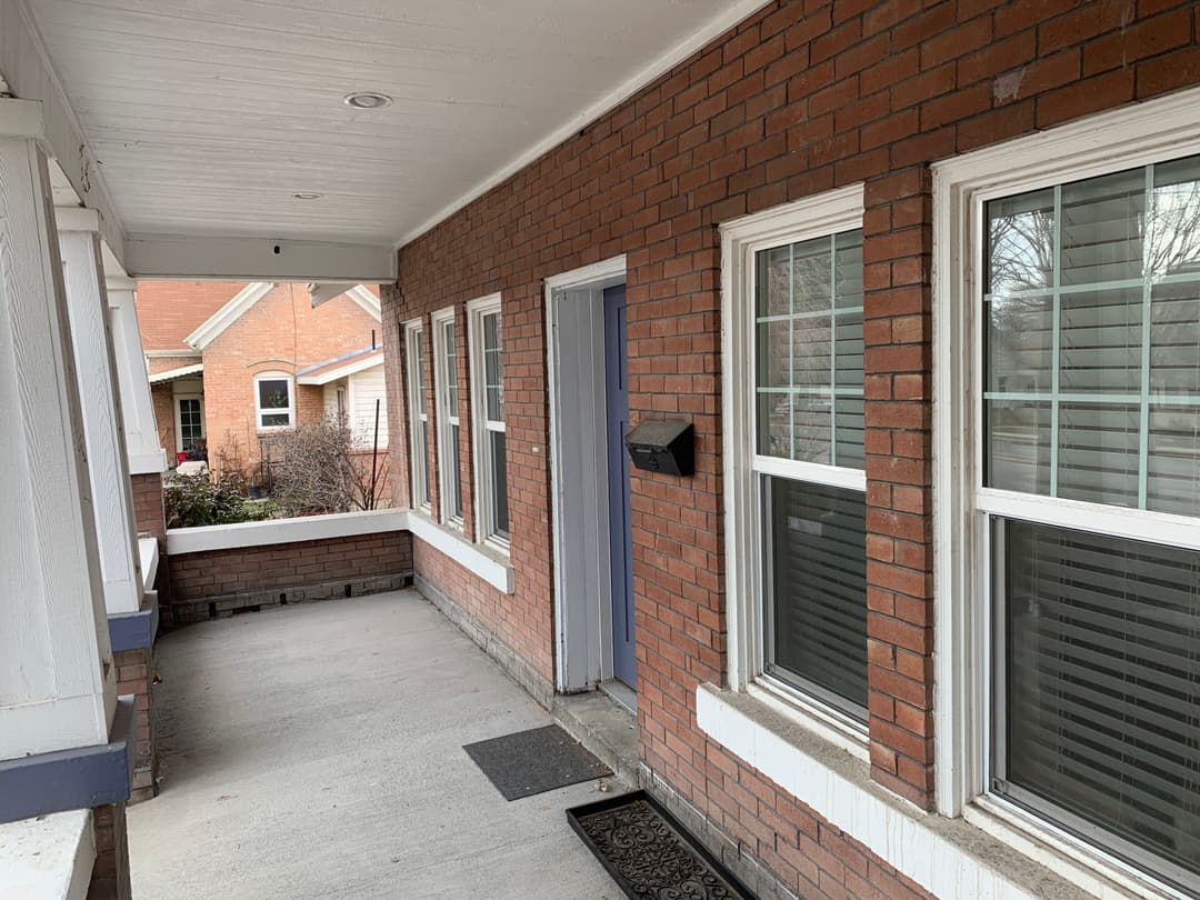 Brick exterior of a home with a porch, front door, windows, and doormat on a sunny day.