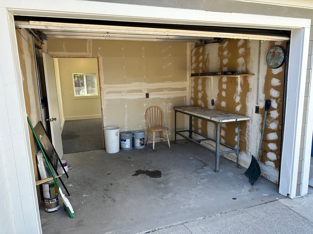 Empty garage space with a workbench, chair, and cleaning supplies, featuring drywall and unfinished walls.
