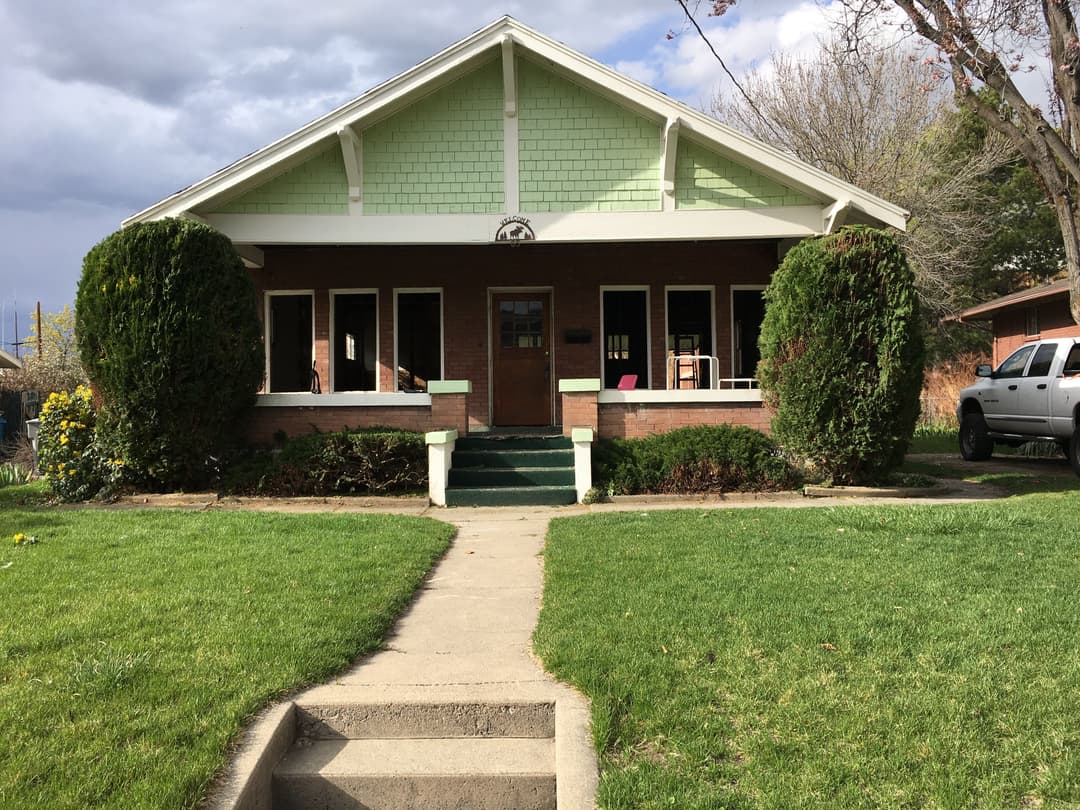 Green and brick bungalow house with flower garden and pathway on a sunny day.