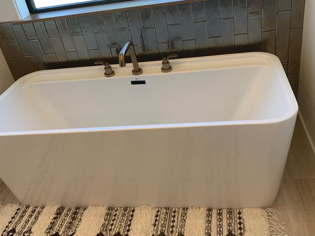 Modern freestanding bathtub with brushed gold faucet, surrounded by a textured tile backsplash.