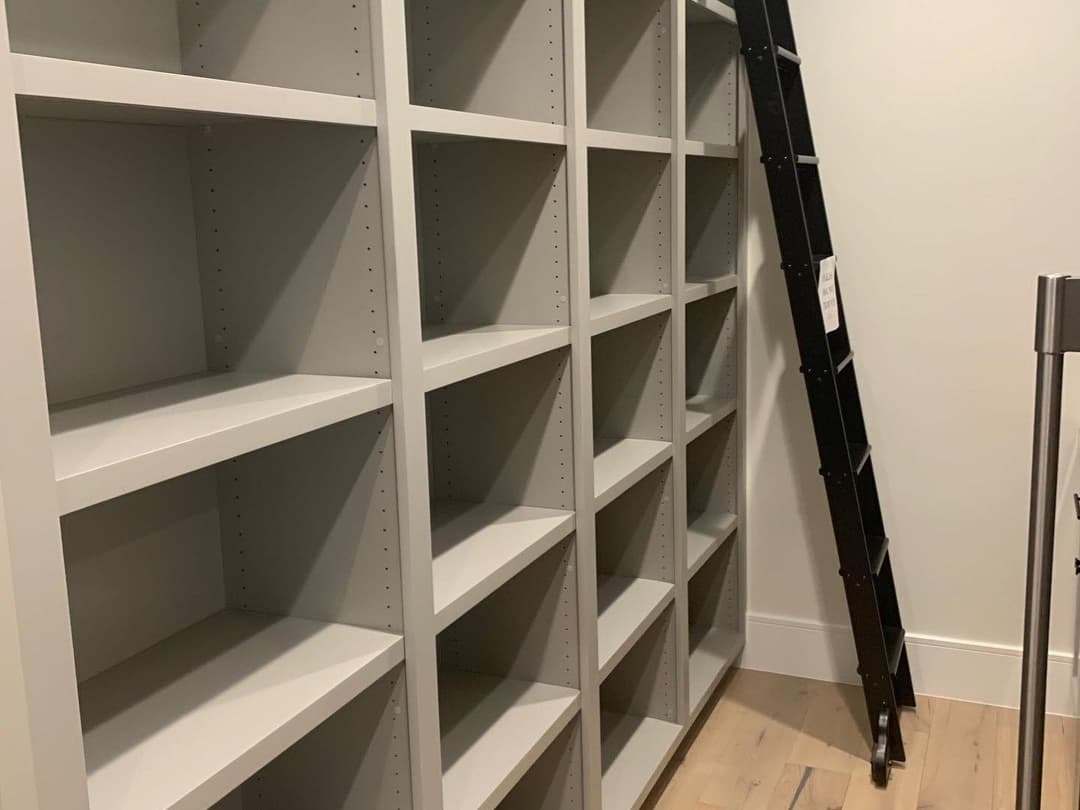 Gray modern bookshelf with empty shelves and a black ladder beside it in a minimalist room.
