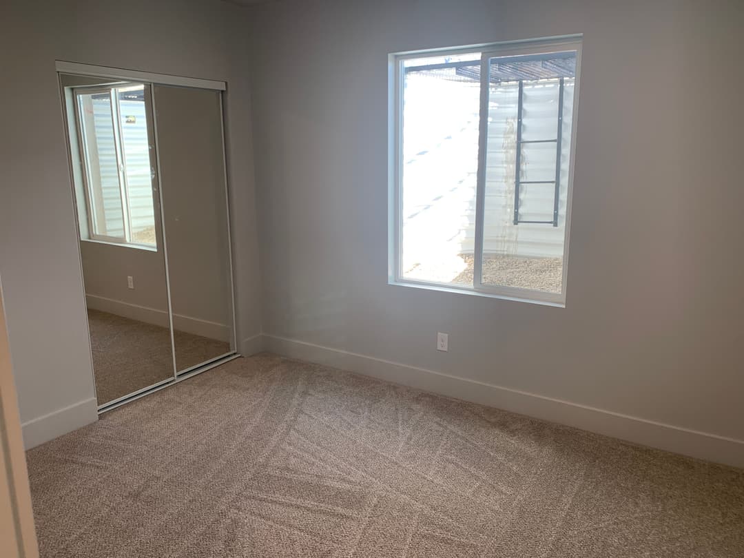 Empty room with carpet, large window, and mirrored closet doors in bright lighting.