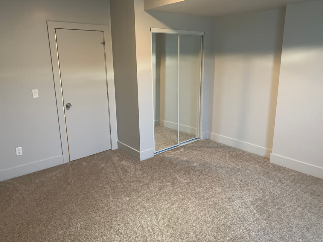 Empty room with gray walls, carpeted floor, closet mirror, and door. Natural light streaming in.