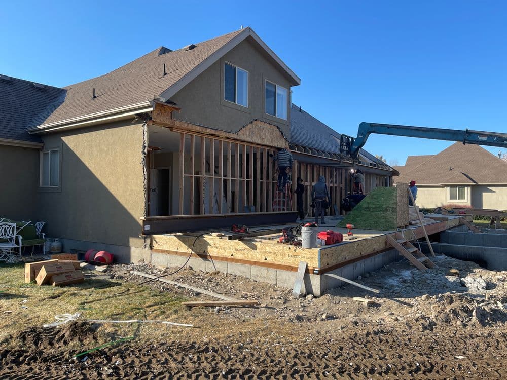 Home renovation in progress with workers framing and using machinery on a sunny day.